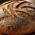 "A freshly baked sourdough bread loaf with a golden crust and airy interior, placed on a rustic wooden board surrounded by a linen cloth, flour-dusted tools, and a jar of sourdough starter."