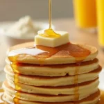 A stack of fluffy golden pancakes made with Krusteaz Pancake Mix, topped with butter and syrup, surrounded by fresh fruits and orange juice.